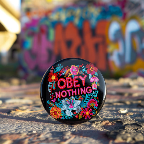 A black round pinback button featuring floral embroidery-style designs and bold red text that reads "Obey Nothing."